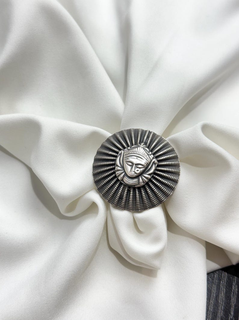 May include: A silver ring with a circular design featuring a stylised face in the centre. The ring is on a white fabric background.