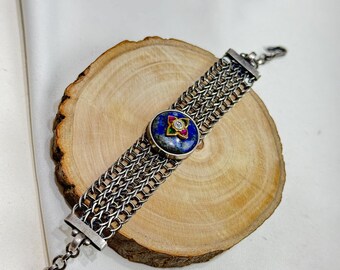 Puede incluir: Un brazalete de cadena de plata con una piedra de lapislázuli azul engastada en un bisel de plata. La piedra está rodeada de piedras más pequeñas de colores. El brazalete está sobre una superficie de madera.