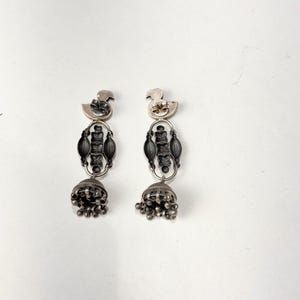 May include: A pair of silver-toned earrings. Each earring features a duck-shaped top, an elongated central design, and a bell-shaped bottom with dangling beads. The earrings are displayed against a white background.