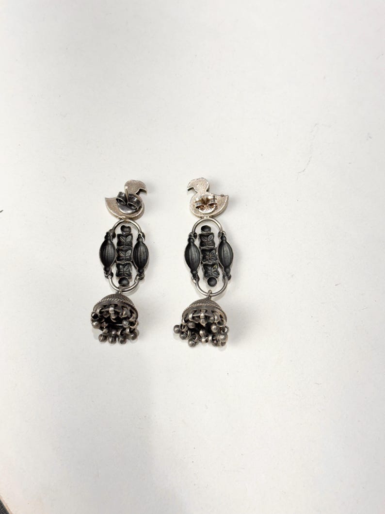 May include: A pair of silver-toned earrings. Each earring features a duck-shaped top, an elongated central design, and a bell-shaped bottom with dangling beads. The earrings are displayed against a white background.