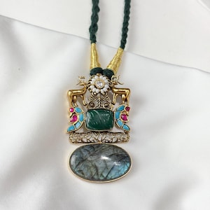 May include: A gold-toned necklace with a green cord. The pendant features a rectangular green stone, an oval gray stone, and a decorative design with deer figures, pink and blue accents, and a white flower.