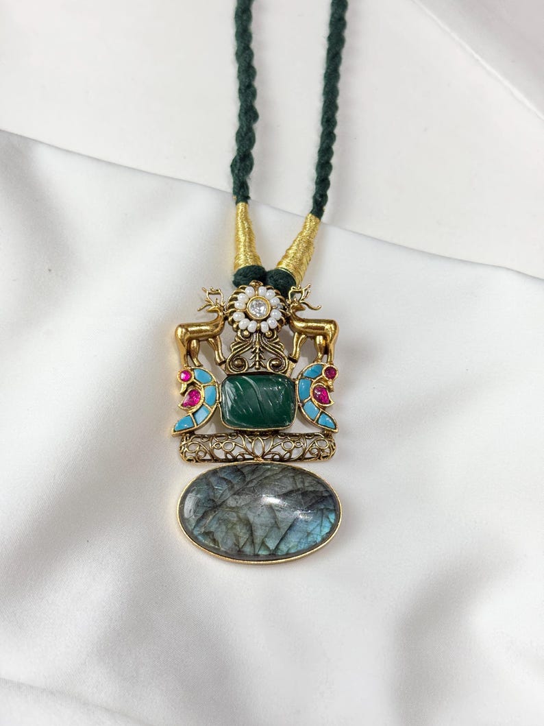 May include: A gold-toned necklace with a green cord. The pendant features a rectangular green stone, an oval gray stone, and a decorative design with deer figures, pink and blue accents, and a white flower.