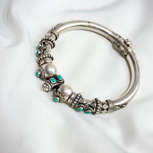 Peut inclure: Un bracelet jonc articulé argenté avec des détails ornés et des accents turquoise. Le bracelet présente des perles et des motifs décoratifs, sur fond blanc. Un bijou, accessoire de mode.