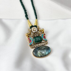 May include: A gold-toned pendant necklace with a dark green cord. The pendant features a large oval-shaped stone, a rectangular green stone, and accents of red, blue, and white. The necklace is displayed on a white surface.