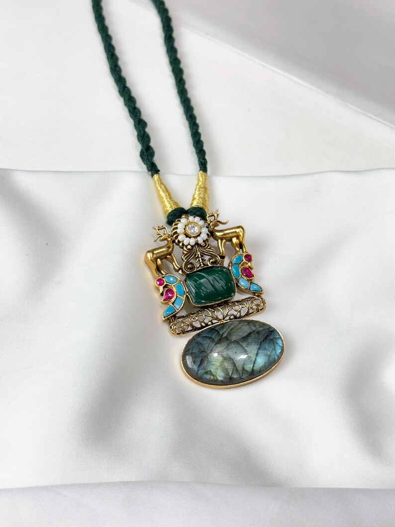 May include: A gold-toned pendant necklace with a dark green cord. The pendant features a large oval-shaped stone, a rectangular green stone, and accents of red, blue, and white. The necklace is displayed on a white surface.