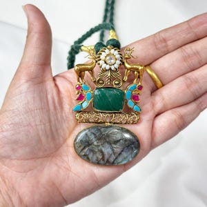 May include: A gold-toned pendant necklace with a green stone center, deer figures, and a large oval stone at the bottom. The necklace has a green cord. The pendant features turquoise and pink accents.