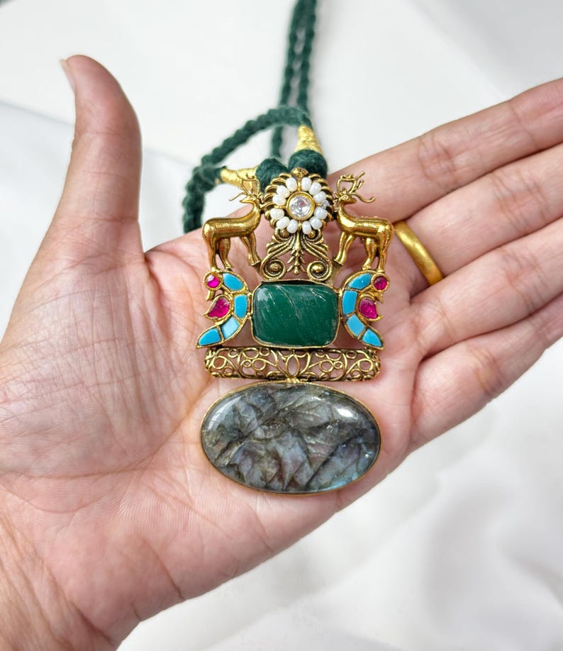 May include: A gold-toned pendant necklace with a green stone center, deer figures, and a large oval stone at the bottom. The necklace has a green cord. The pendant features turquoise and pink accents.