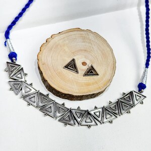Puede incluir: Un collar de plata con un cordón azul y un colgante de plata hecho de múltiples formas triangulares. El colgante está sobre una superficie de madera con dos pequeños pendientes triangulares de plata.