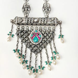 May include: Silver-toned necklace with turquoise and pink gemstone accents. The necklace features a central pendant with a turquoise stone surrounded by pink and white detailing. Dangling chains hold white teardrop-shaped pearls and green beads. The necklace has two matching earrings.