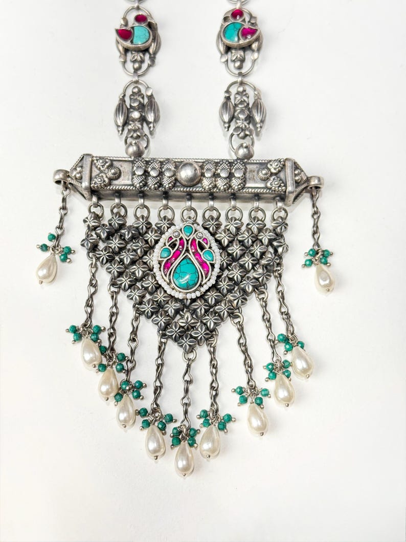 May include: Silver-toned necklace with turquoise and pink gemstone accents. The necklace features a central pendant with a turquoise stone surrounded by pink and white detailing. Dangling chains hold white teardrop-shaped pearls and green beads. The necklace has two matching earrings.