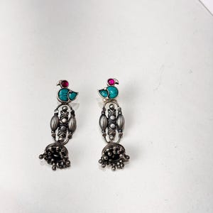 May include: A pair of silver-toned dangle earrings. Each earring features a turquoise bird design with a red accent stone. The earrings have an ornate, elongated design with a bell-shaped bottom.