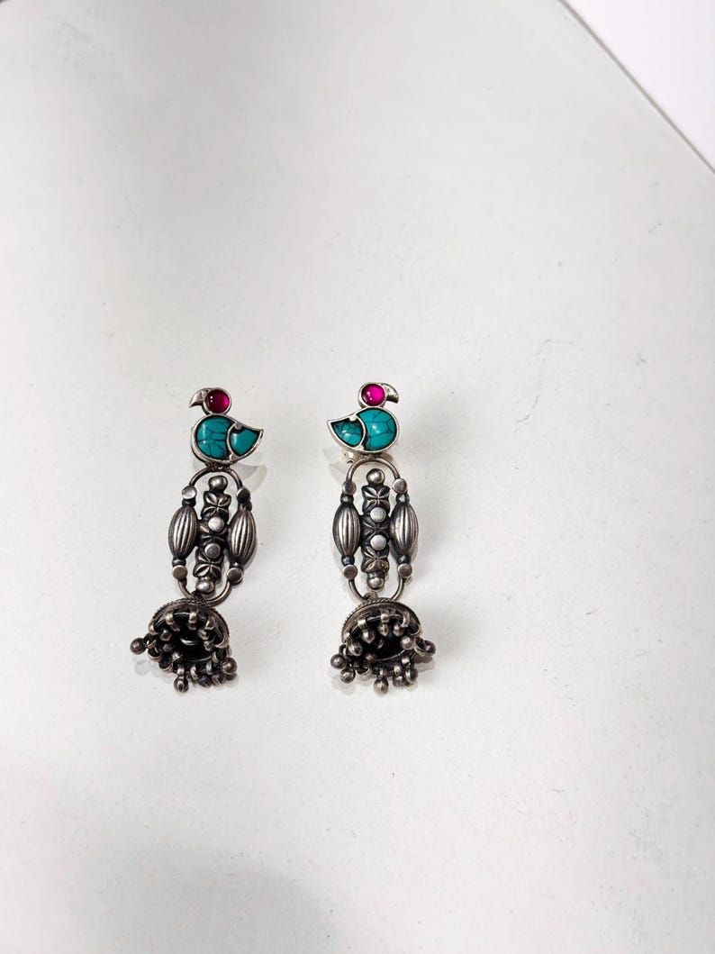 May include: A pair of silver-toned dangle earrings. Each earring features a turquoise bird design with a red accent stone. The earrings have an ornate, elongated design with a bell-shaped bottom.