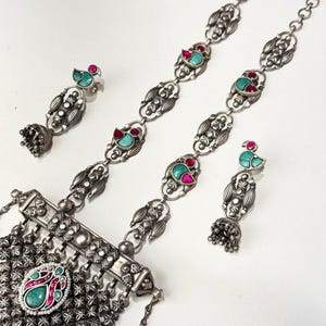 May include: A silver-toned jewelry set featuring a necklace and two pairs of earrings. The necklace has a central pendant with a turquoise and pink stone, and the earrings and necklace have turquoise and red accents.