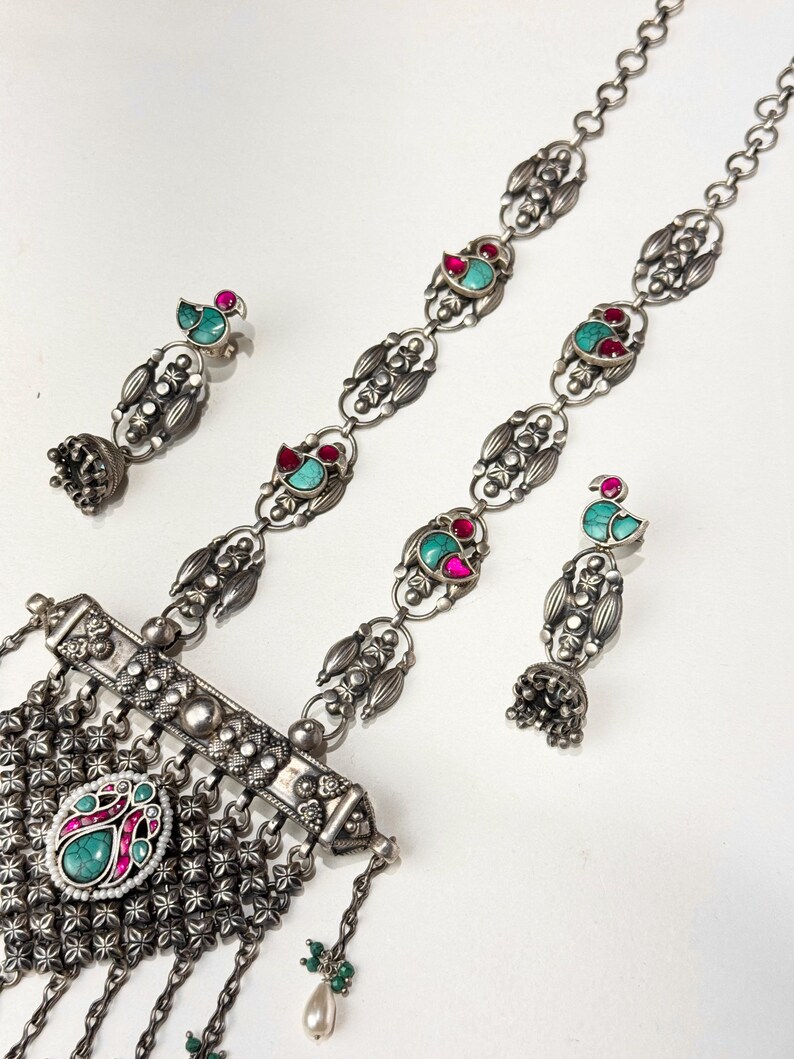 May include: A silver-toned jewelry set featuring a necklace and two pairs of earrings. The necklace has a central pendant with a turquoise and pink stone, and the earrings and necklace have turquoise and red accents.