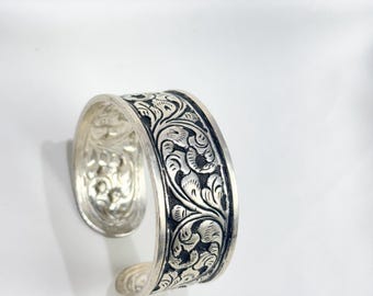 May include: A silver cuff bracelet with an intricate floral design. The bracelet is open and has a smooth, polished finish.