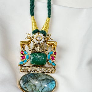 May include: A gold-toned pendant necklace with a green cord. The pendant features a green square stone, gold deer figures, and a large oval stone with blue and green hues. Accents include red and turquoise stones.