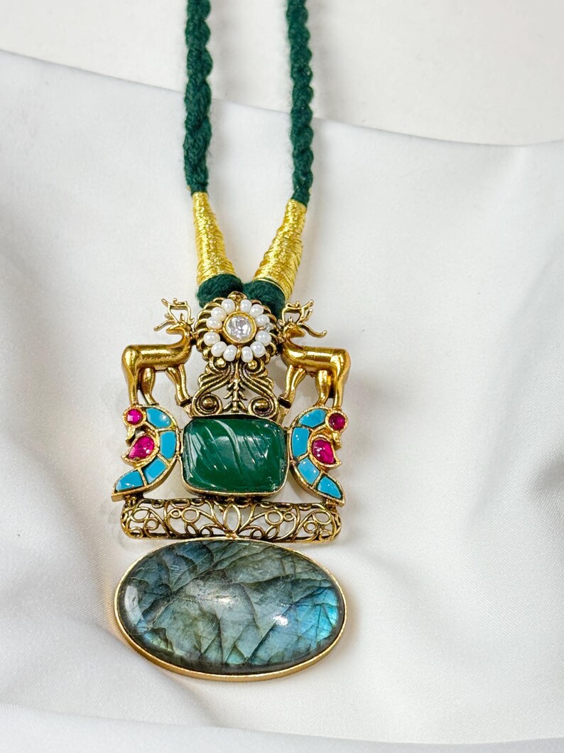 May include: A gold-toned pendant necklace with a green cord. The pendant features a green square stone, gold deer figures, and a large oval stone with blue and green hues. Accents include red and turquoise stones.