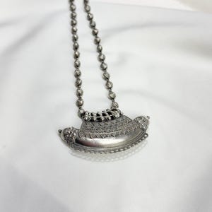 May include: A silver-toned necklace with a pendant. The pendant has an ornate, boat-like design with intricate detailing. The necklace chain is composed of small, round beads. The jewelry is displayed on a white background.