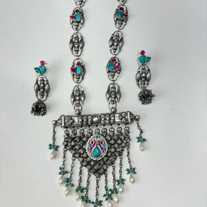 May include: A silver-toned necklace and earring set. The necklace features a central pendant with turquoise and pink accents, and dangling pearl-like beads. The earrings and necklace have matching turquoise and pink accents.