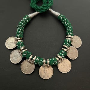 May include: A green and silver necklace with a tassel. The necklace is made of a braided cord with silver coins attached. The coins have a design of a person's head. The necklace weighs 74 grams.