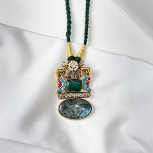May include: A gold-toned pendant necklace with a green cord. The pendant features a large oval-shaped, iridescent stone at the bottom, a square green stone, and decorative elements with red and turquoise accents. The necklace has a dark green cord.