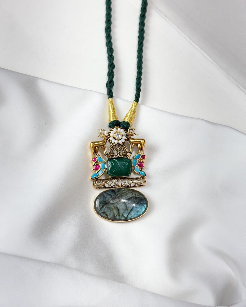 May include: A gold-toned pendant necklace with a green cord. The pendant features a large oval-shaped, iridescent stone at the bottom, a square green stone, and decorative elements with red and turquoise accents. The necklace has a dark green cord.