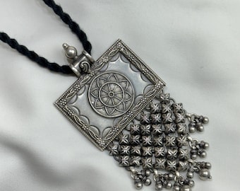 May include: A silver pendant necklace with a square-shaped pendant featuring an intricate floral design and a tassel of small silver beads. The necklace is strung on a black cord.
