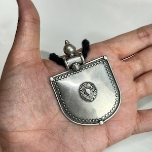May include: A silver-toned pendant necklace with a shield-shaped design. The pendant features a central floral motif and a beaded border. The necklace has a black cord. The pendant is approximately 2 inches in length.