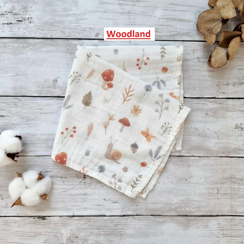 Pu&ograve; includere: Un bavaglino in mussola di cotone bianco con una stampa a tema bosco che presenta funghi, foglie, ghiande e una lumaca. Il testo "Woodland" &egrave; stampato in rosso nella parte superiore dell'immagine.