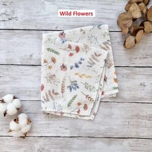 Pu&ograve; includere: Un quadrato di tessuto bianco con un motivo di fiori selvatici e foglie colorate. Il tessuto &egrave; piegato a met&agrave; e il testo "Wild Flowers" &egrave; stampato in rosso nella parte superiore dell'immagine.