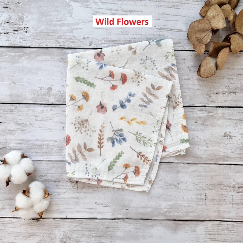 Pu&ograve; includere: Un quadrato di tessuto bianco con un motivo di fiori selvatici e foglie colorate. Il tessuto &egrave; piegato a met&agrave; e il testo "Wild Flowers" &egrave; stampato in rosso nella parte superiore dell'immagine.