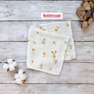 Pu&ograve; includere: Un quadrato di tessuto bianco con un motivo di fiori gialli. Il tessuto &egrave; piegato a met&agrave; e il testo "Buttercups" &egrave; stampato su un'etichetta bianca.