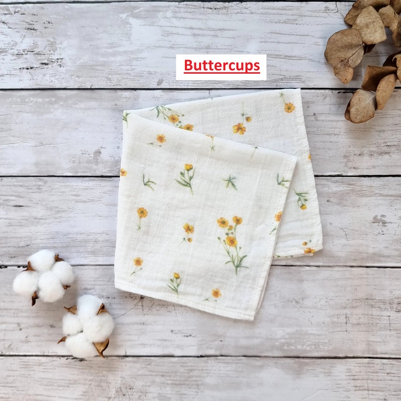 Pu&ograve; includere: Un quadrato di tessuto bianco con un motivo di fiori gialli. Il tessuto &egrave; piegato a met&agrave; e il testo "Buttercups" &egrave; stampato su un'etichetta bianca.