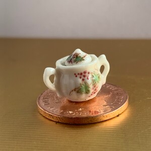 Puede incluir: Un azucarero en miniatura de porcelana blanca con tapa, que presenta un diseño floral de hojas verdes y bayas rojas. El azucarero está sobre una moneda de color cobre.