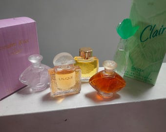 May include: Five miniature perfume bottles, including a clear bottle with a floral design, a clear bottle with a gold top, a clear bottle with a gold top and a floral design, a clear bottle with a gold top and a floral design, and a green bottle with a floral design. The bottles are on a white surface. The bottles are labeled "Lalique" and "Claire".