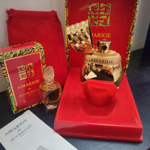 May include: A gold bottle of Amarigé perfume by Givenchy, with a gold cap and a red box. The box has a colourful pattern and the words "Amarigé de Givenchy" on it. The bottle is sitting on a red tray with a red velvet bag in the background.