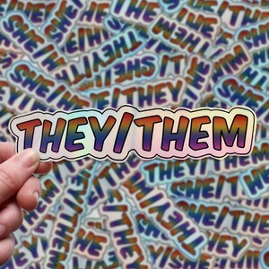 May include: A rainbow holographic sticker with the text "THEY/THEM" in bold, white letters. The sticker is being held by a hand against a background of many similar stickers with different pronoun combinations.
