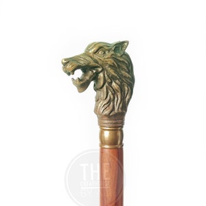 Pode incluir: Uma bengala de madeira com uma pega de cabe&ccedil;a de lobo de bronze detalhada. A boca do lobo est&aacute; aberta e os seus olhos est&atilde;o alerta. A bengala &eacute; castanha e tem um acabamento liso.