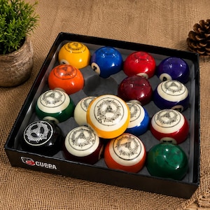 Op de afbeelding: Een zwarte doos met een set van zestien biljartballen, elk voorzien van een uniek cirkelvormig embleem op het oppervlak. De ballen zijn in een raster gerangschikt en vertonen verschillende effen kleuren, waaronder geel, blauw, rood, oranje, groen en zwart.