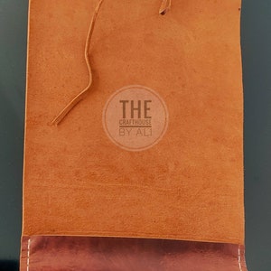 May include: A brown leather journal with a flap closure and a brown leather strap. The journal has a pocket on the front and the text "THE Crafthouse BY ALI" is printed on the front.