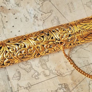 May include: A gold metal scroll tube with intricate floral designs. The tube has a chain attached to a decorative cap. The tube is open and a rolled piece of paper is partially visible.