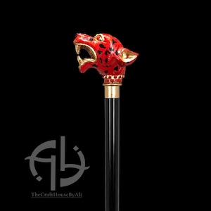 Könnte beinhalten: Ein Gehstock mit einem skulptierten Leopardenkopf-Griff, der in leuchtendem Rot mit schwarzen Flecken bemalt ist und goldfarbene Akzente an Maul und Ohren aufweist. Der Griff sitzt auf einem glatten, schwarzen Schaft, verbunden durch eine goldfarbene Metallmanschette.