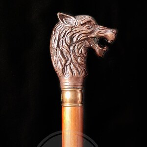 Pode incluir: Uma bengala de madeira com uma pega de cabe&ccedil;a de lobo de cobre detalhada. A boca do lobo est&aacute; aberta e os seus olhos est&atilde;o alerta.