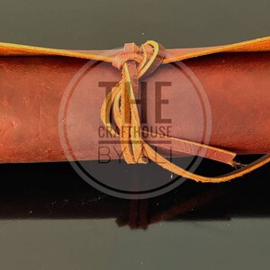 May include: A rolled-up piece of brown leather with a tan leather strap tied around it. The strap is tied in a knot and the ends are frayed. The leather is textured and has a rustic look. The text "THE Crafthouse BY" is visible on the strap.