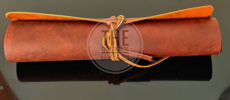 May include: A rolled-up piece of brown leather with a tan leather strap tied around it. The strap is tied in a knot and the ends are frayed. The leather is textured and has a rustic look. The text "THE Crafthouse BY" is visible on the strap.
