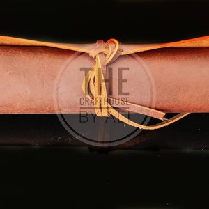 May include: A brown leather roll with a tan leather strap tied around it. The strap is tied in a knot and the ends are left loose. The roll is on a black background. The text "THE Crafthouse BY ALI" is visible on the strap.