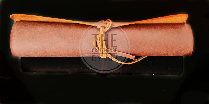 May include: A brown leather roll with a tan leather strap tied around it. The strap is tied in a knot and the ends are left loose. The roll is on a black background. The text "THE Crafthouse BY ALI" is visible on the strap.