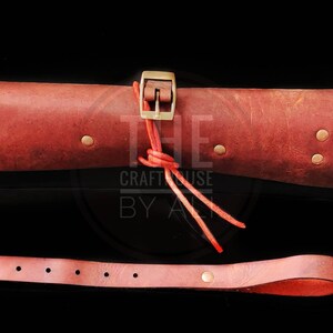 Op de afbeelding: Een opgerolde leren draagtas met een messing gesp en een rode leren riem. De tas is gesloten met een rode leren riem en vastgebonden met een rood koord. De tas bevindt zich op een zwarte achtergrond. De tekst "THE CRAFT HOUSE BY ALI" is zichtbaar in de afbeelding.