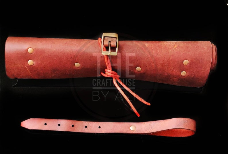 Op de afbeelding: Een opgerolde leren draagtas met een messing gesp en een rode leren riem. De tas is gesloten met een rode leren riem en vastgebonden met een rood koord. De tas bevindt zich op een zwarte achtergrond. De tekst "THE CRAFT HOUSE BY ALI" is zichtbaar in de afbeelding.