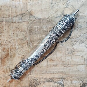 Pu&ograve; includere: Un astuccio argentato per pergamene con intricati motivi floreali e geometrici. La custodia cilindrica ha tappi decorativi e una catena. Lo sfondo &egrave; una mappa vintage.